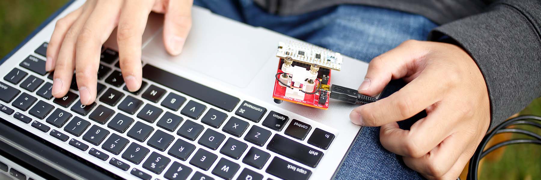 A close-up of a student's hands as they work with a circuit board, USB cable and a laptop